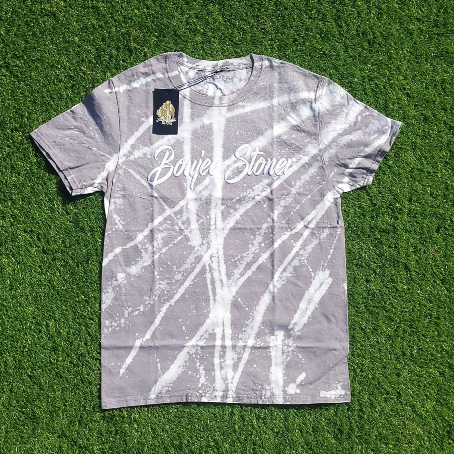 A stylish Boujee Stoner Bleached Tee in a cool grey "Stonewash Grey" color with a distinctive white bleach pattern, showcased against a green grassy background.