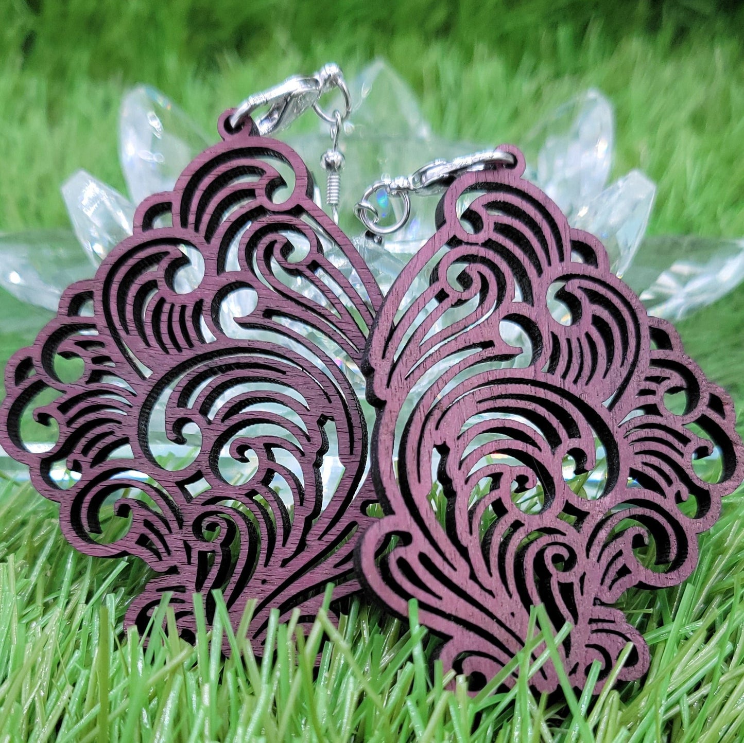 A pair of intricately designed Boujee Stoner Vapor Earrings crafted from purple heart wood, lying on a bed of green grass.