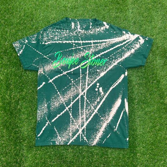 A relaxed-fit Boujee Stoner Bleached Tee in a deep forest green with a unique white splatter pattern, laid flat on a grassy background.