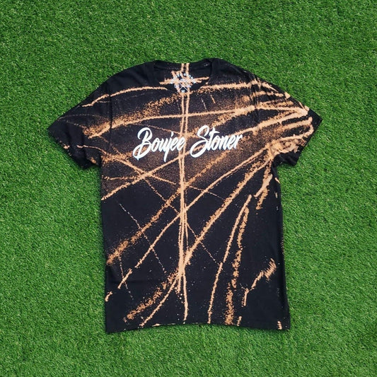 A black Boujee Stoner Bleached Tee with a striking white bleach splatter effect, laid flat against a green grass backdrop.