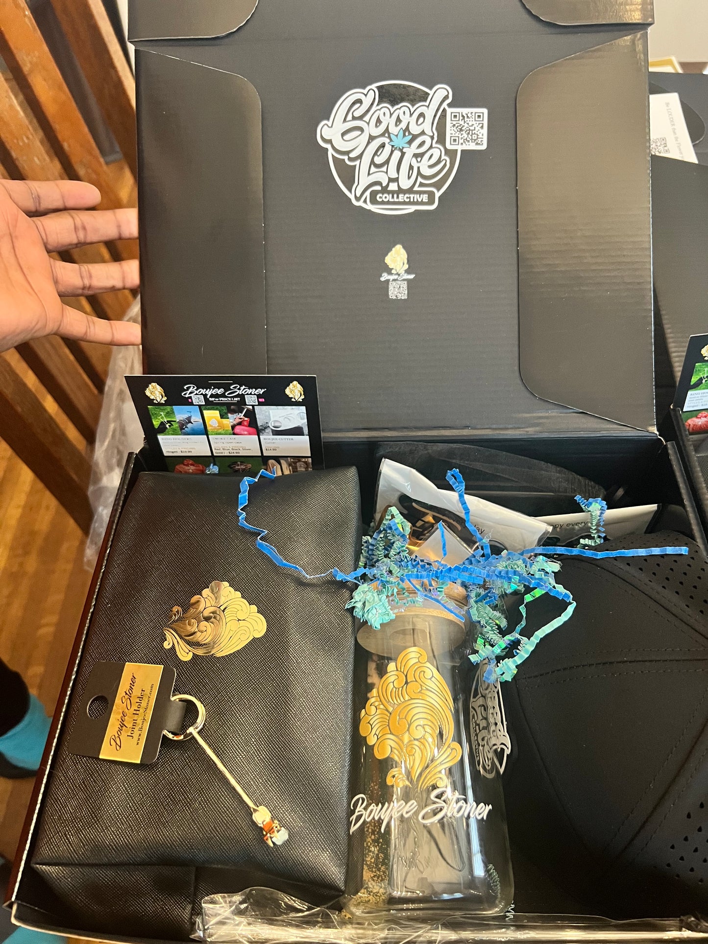 Hand opening a luxurious black Boujee Box filled with gold and blue smoking accessories, including a grinder, rolling papers, and a sleek black pouch.
